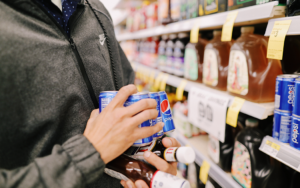 Person in grey zip-up sweater in a grocery store holding a can of Pepsi and other products | ChefsBest blog "Private Brands: Keeping up with National Legacy Brands"